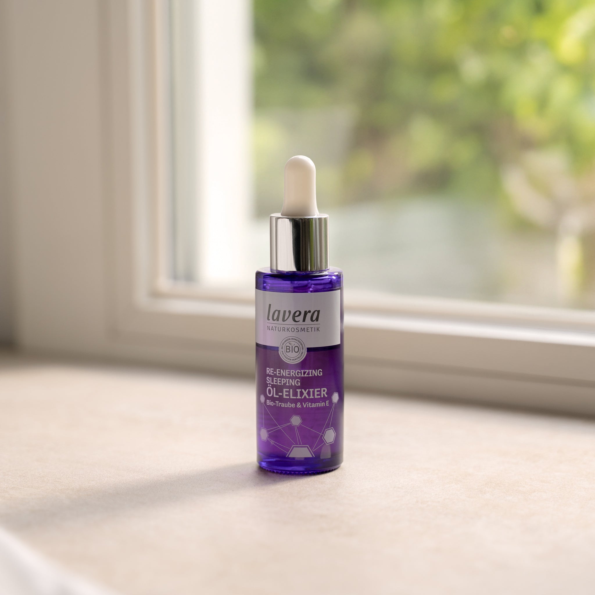 Fioletowa szklana butelka z pipetą Re-Energizing Sleeping Oil Elixir lavera na parapecie w miękkim świetle; w tle rozmyta zieleń