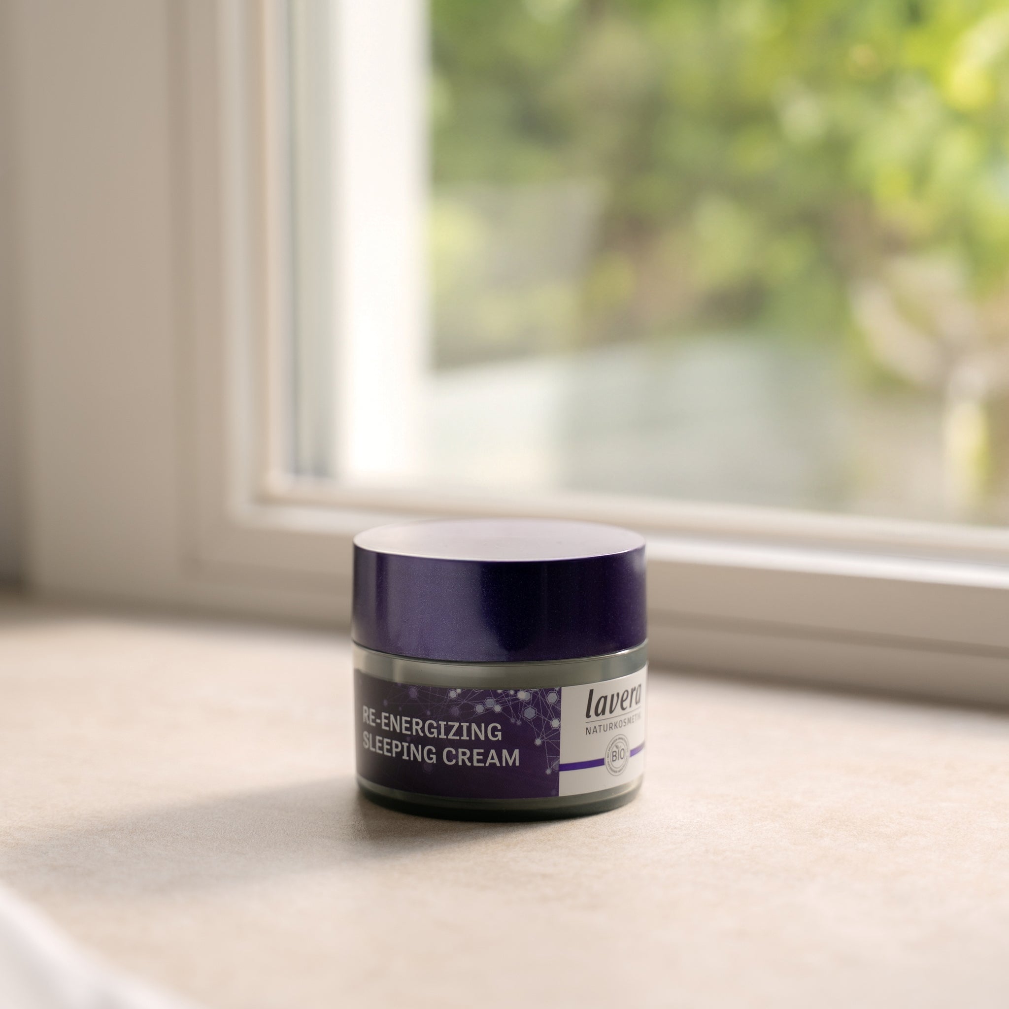 Okrągły słoiczek z fioletową pokrywką Re-Energizing Sleeping Cream lavera na parapecie w miękkim świetle; rozmyta zieleń w tle