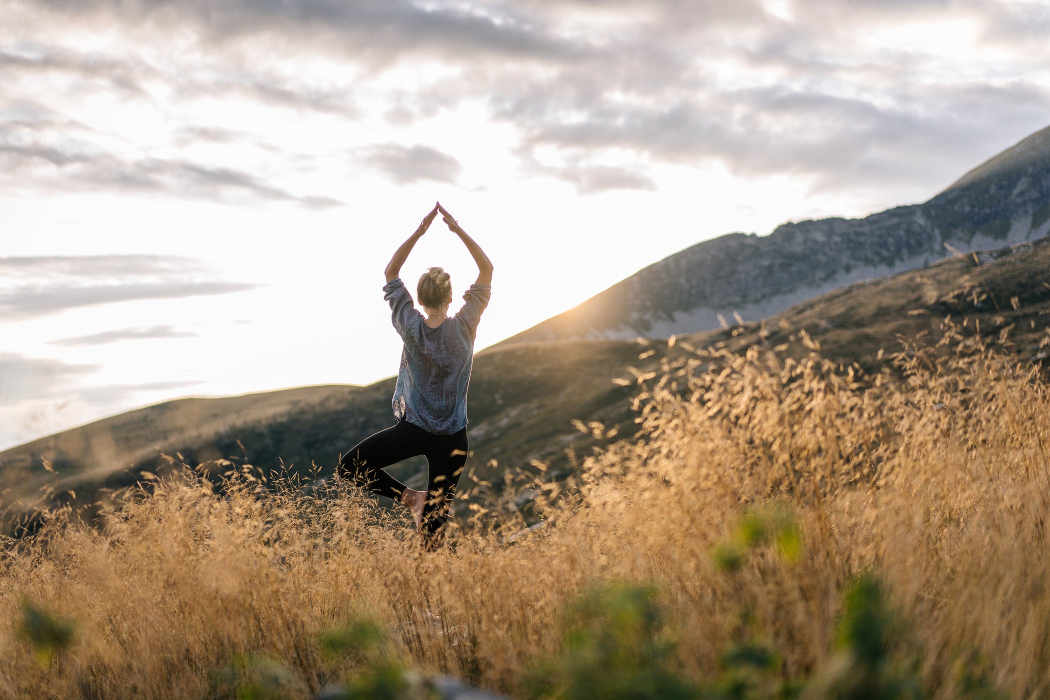 Frau steht mit dem Rücken zur Kamera inmitten einer Wiese in den Bergen und macht eine Yogaposition