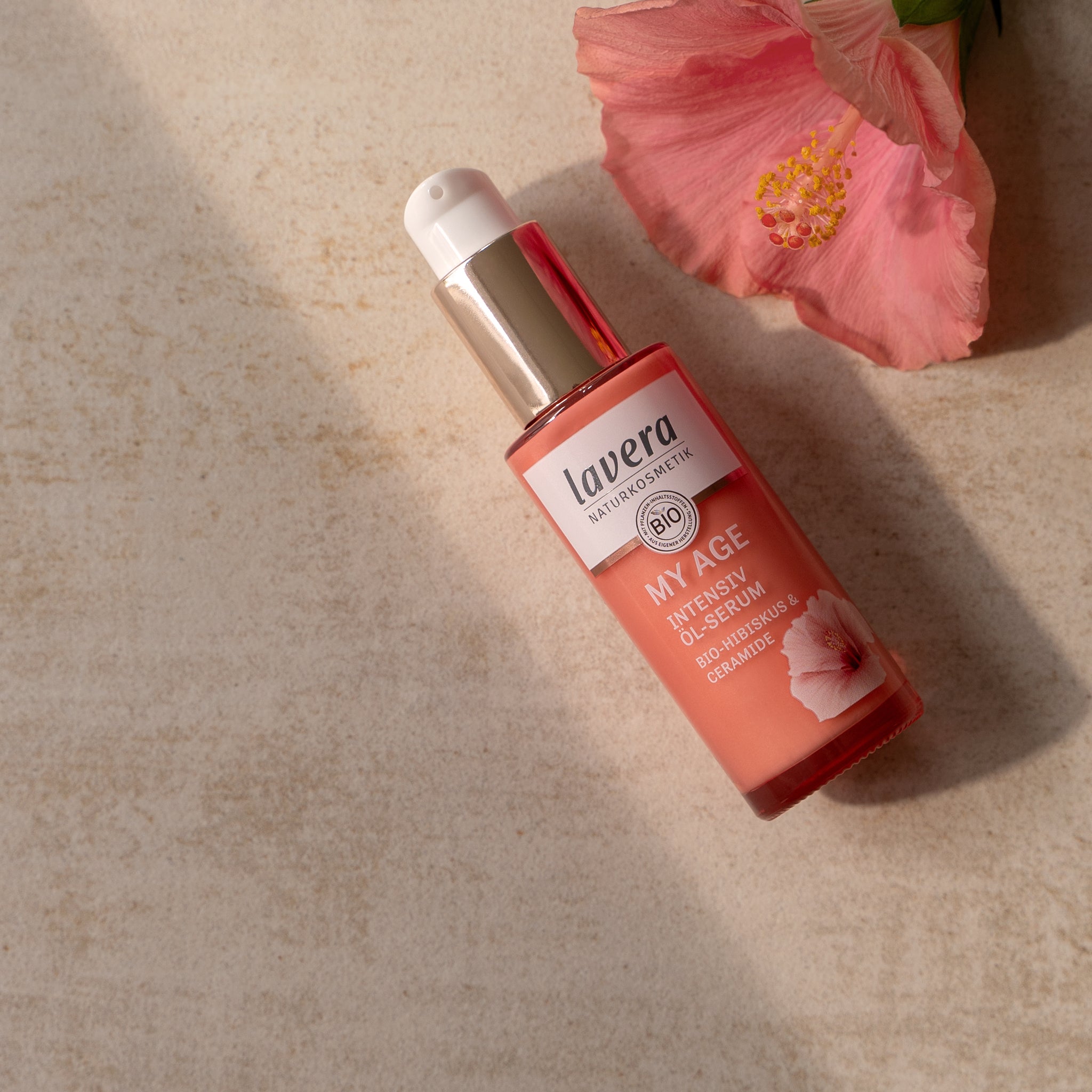 lavera MY AGE intensywne olejowe serum z hibiskusem bio i ceramidami, ustawione obok różowego kwiatu hibiskusa na jasnej powierzchni kamiennej