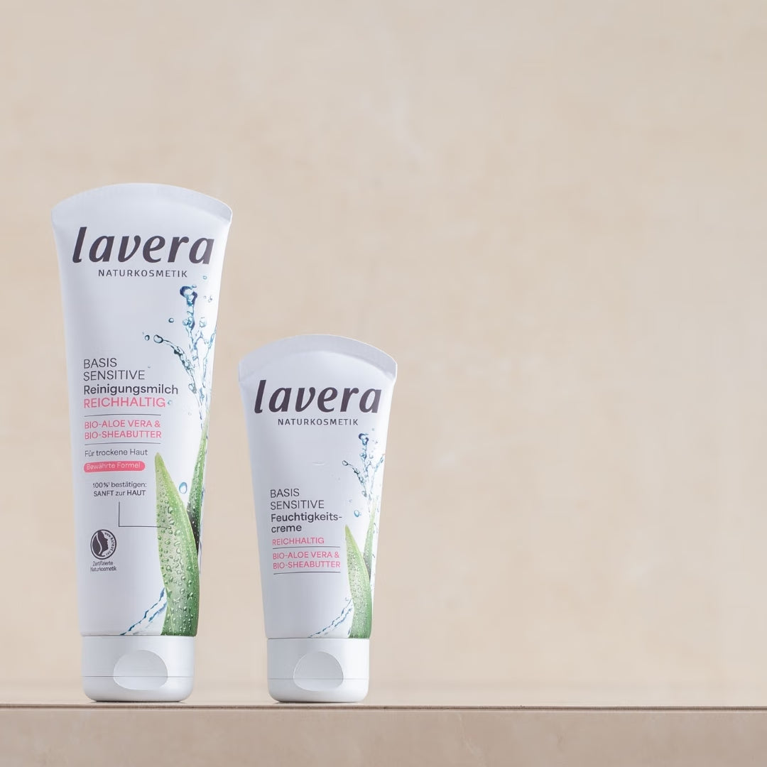 Zwei lavera Basis Sensitive Pflegeprodukte – reichhaltige Reinigungsmilch und Feuchtigkeitscreme – stehen auf einem beigefarbenen Podest.