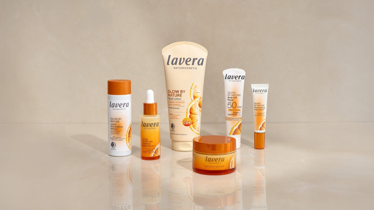Sechs lavera Glow by Nature Pflegeprodukte stehen auf einer spiegelnden Oberfläche vor einem hellen, beigefarbenen Hintergrund.