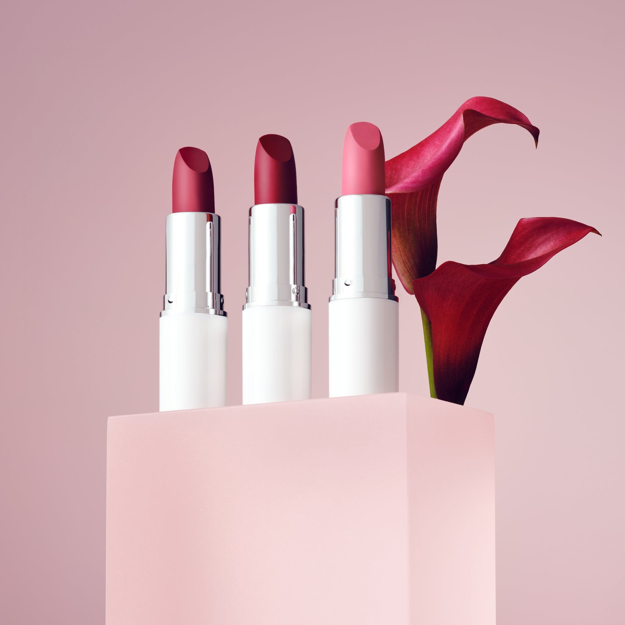 Trzy szminki w odcieniach czerwieni ozdobnie ustawione na różowym tle z kwiatem