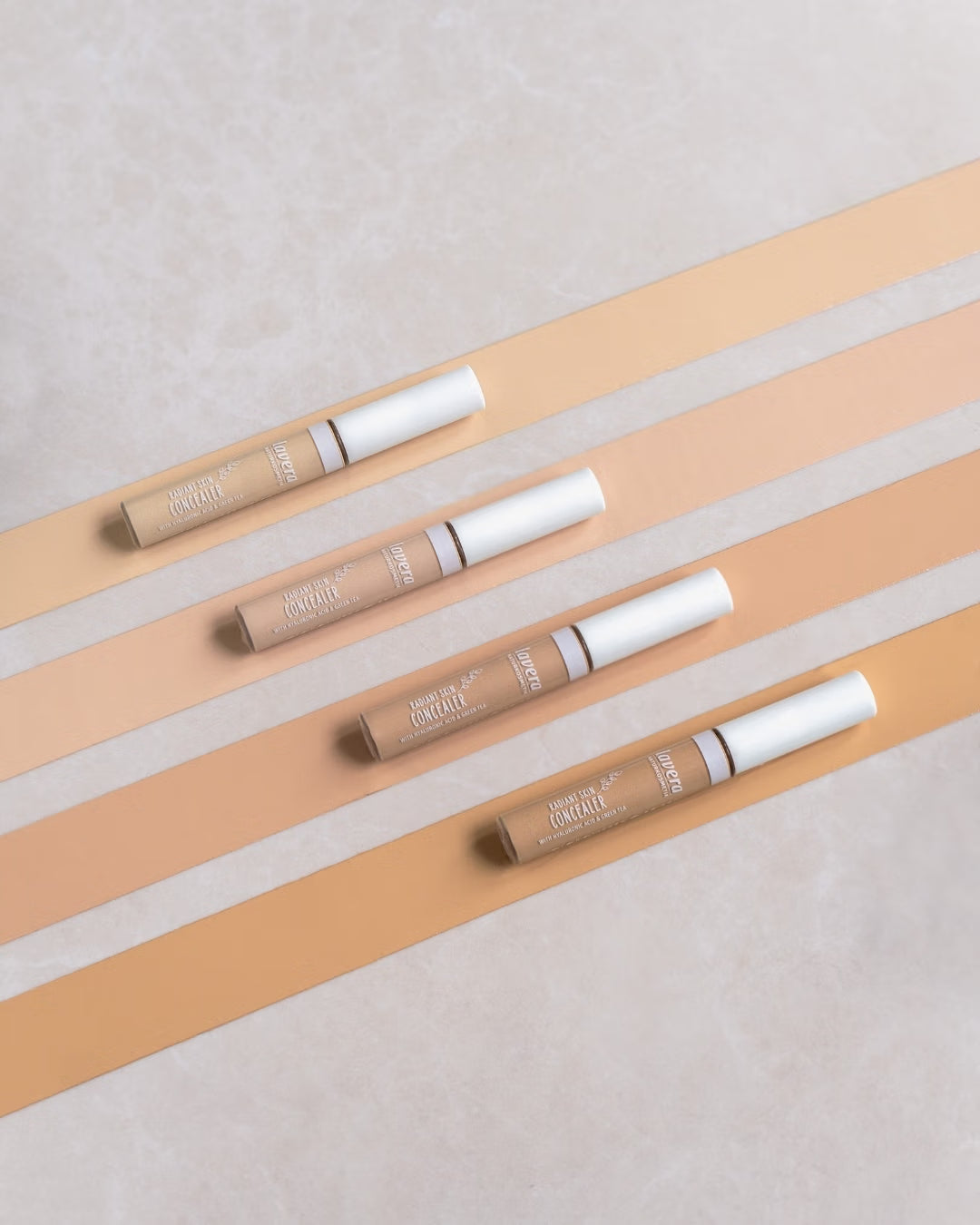 Cztery korektory lavera Radiant Skin Concealer w różnych odcieniach na beżowym wzorze w paski.