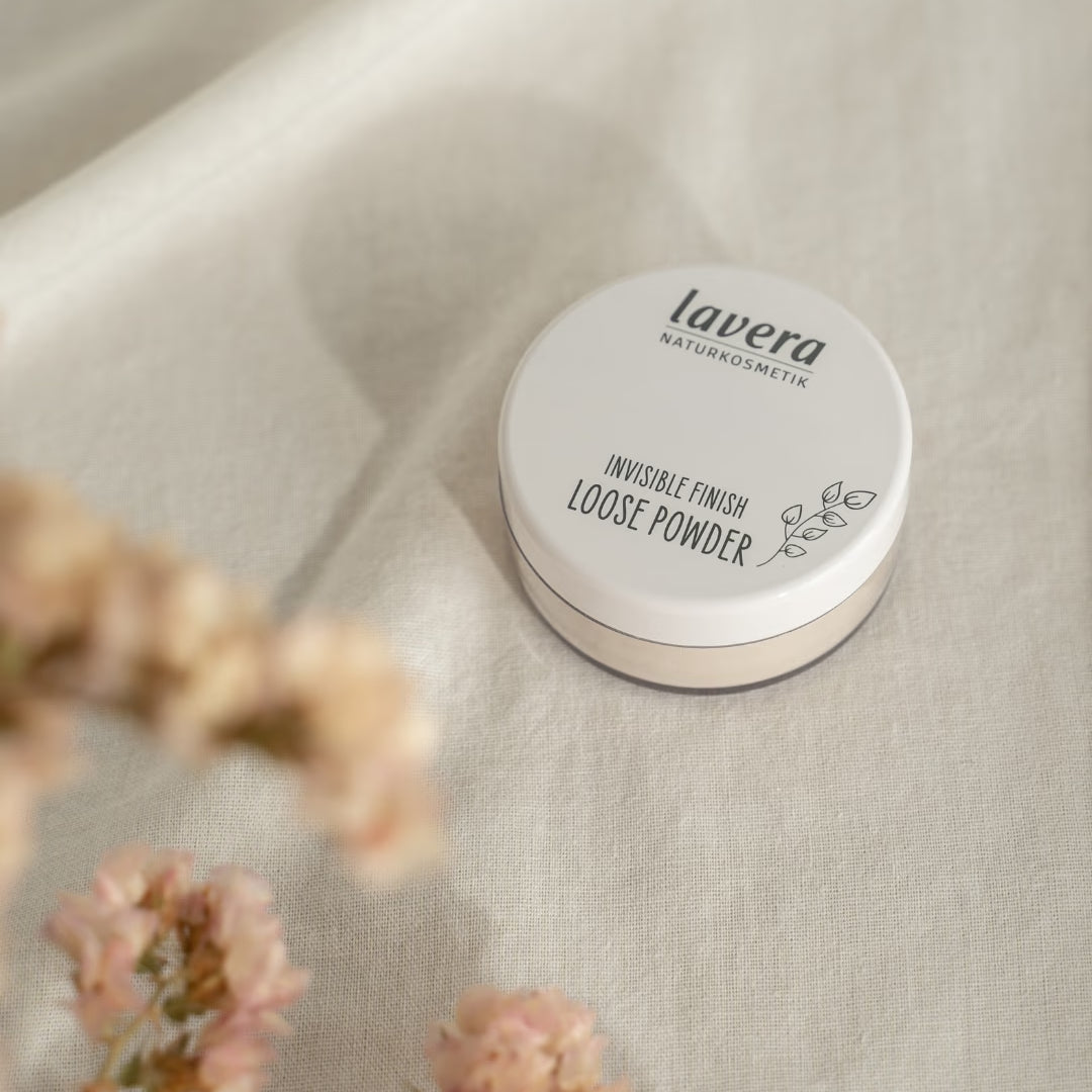 Puder sypki lavera Invisible Finish leży na jasnym lnianym materiale, otoczony rozmytymi różowymi kwiatami.