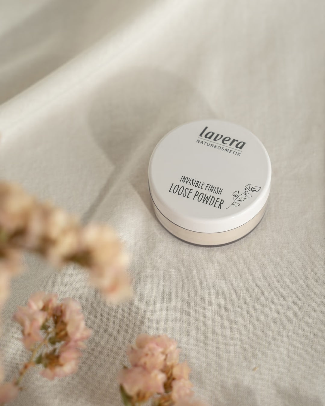 Puder sypki lavera Invisible Finish leży na jasnym lnianym materiale, otoczony rozmytymi różowymi kwiatami.