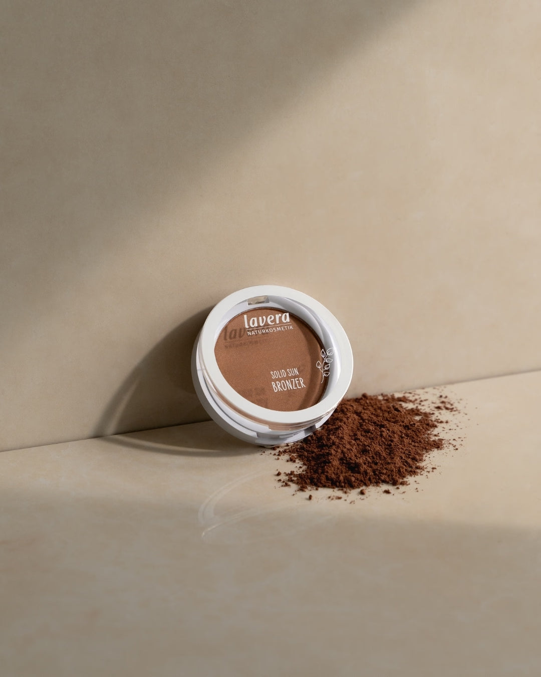 lavera Solid Sun Bronzer liegt auf einer beigefarbenen Oberfläche neben verstreutem Bronzerpuder.