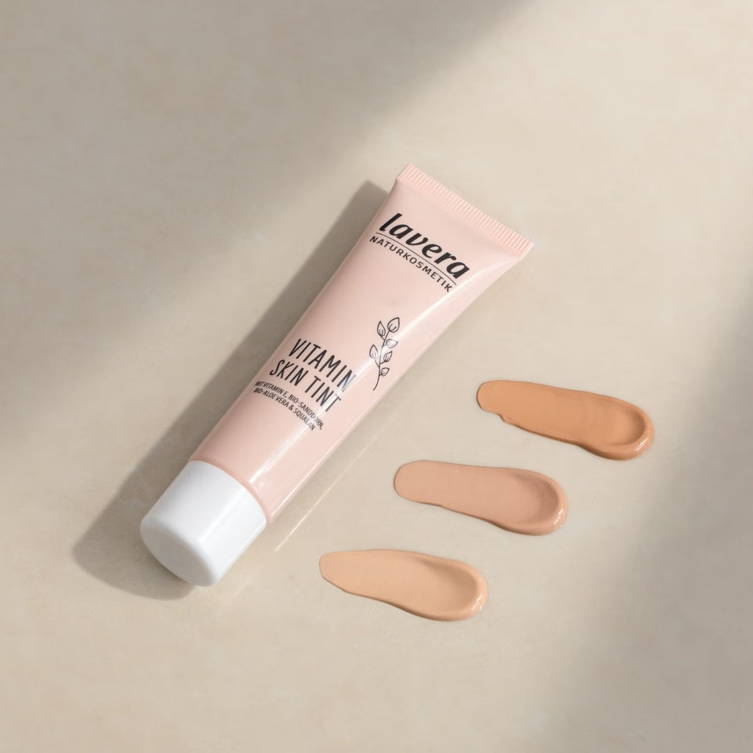 Tuba lavera Vitamin Skin Tint leży obok trzech próbek kolorów o różnej jasności na beżowej powierzchni.