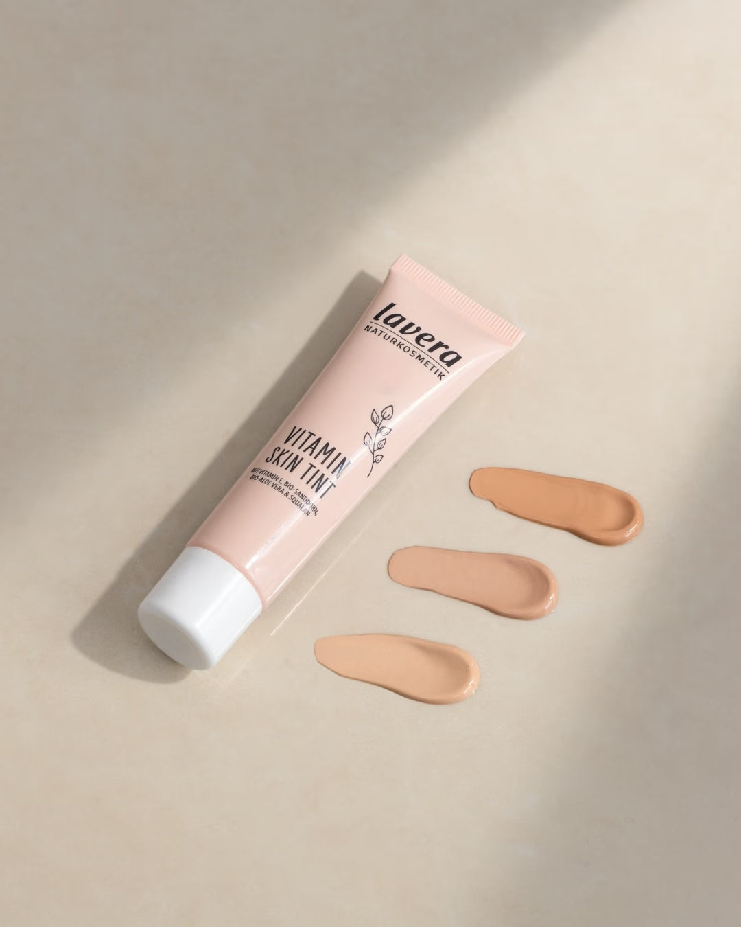 Tuba lavera Vitamin Skin Tint leży obok trzech próbek kolorów o różnej jasności na beżowej powierzchni.