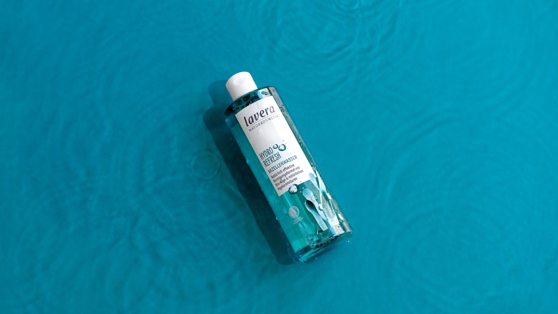 Eine Flasche lavera Hydro Refresh Mizellenwasser liegt auf einer leuchtend türkisblauen, strukturierten Oberfläche im Sonnenlicht.