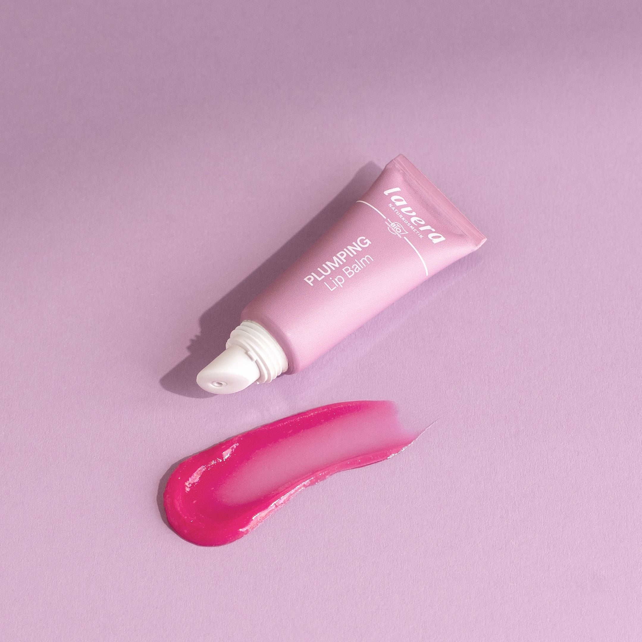 Lavera Plumping Lip Balm in rosa Tube mit aufgetragener pinker Textur auf violettem Hintergrund.