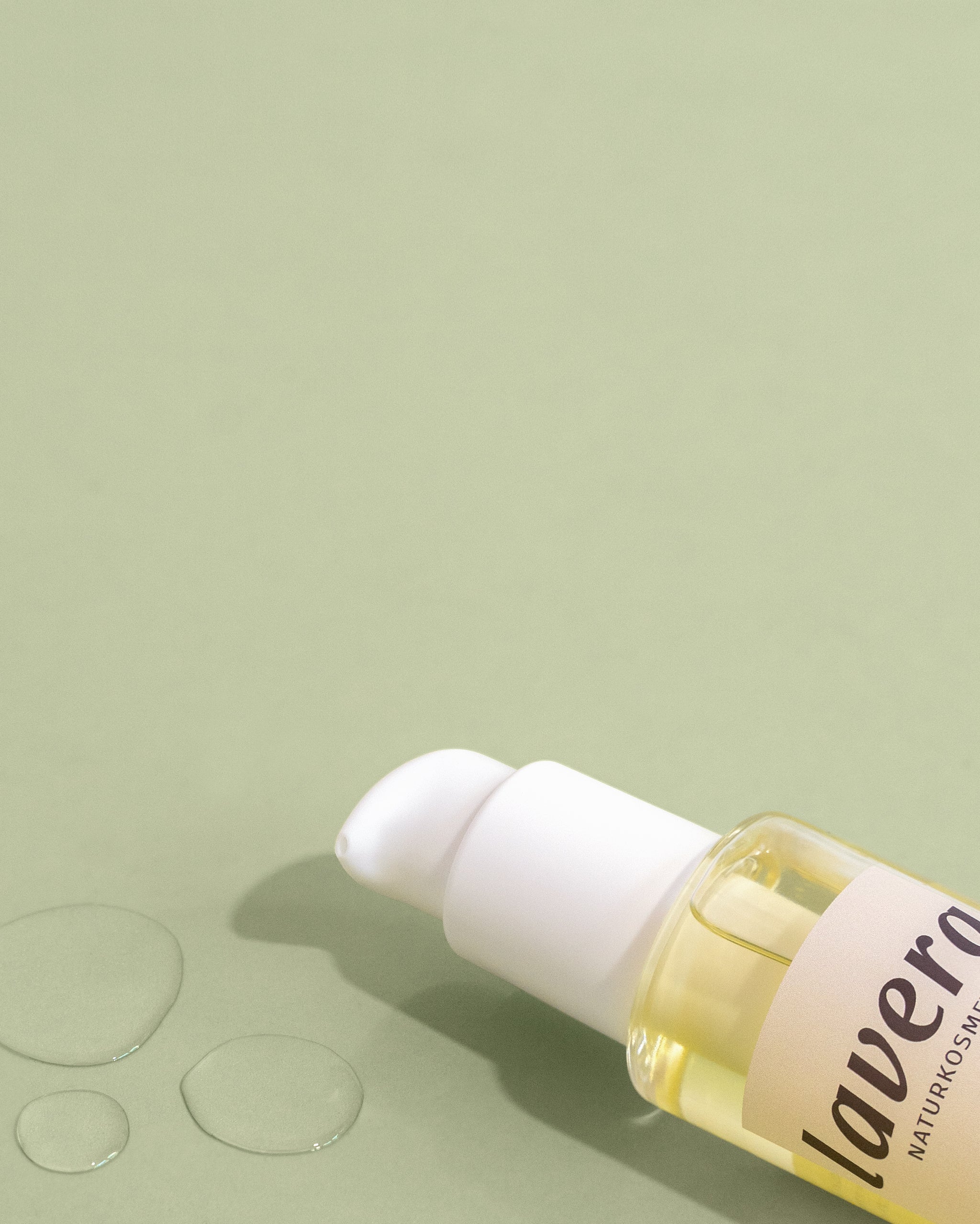 Flasche Lavera Naturkosmetik Baby-Pflegeöl mit Pumpspender, liegend auf grünem Hintergrund mit Öltropfen daneben.