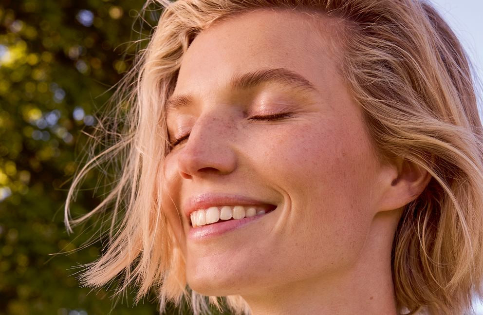 Eine blonde junge Frau, die draußen mit geschlossenen Augen lächelt. Goldfarbene Sonnenstrahlen erhellen das Gesicht von links.